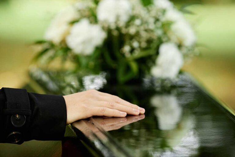 A middle-aged man and woman sit quietly on a park bench, holding hands and looking thoughtfully at a framed photograph.
